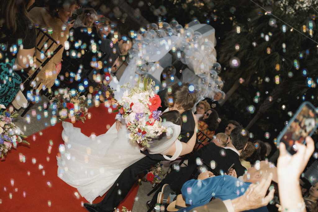 groom dips and kisses bride at the end of the ceremony aisle while bubbles fill the air